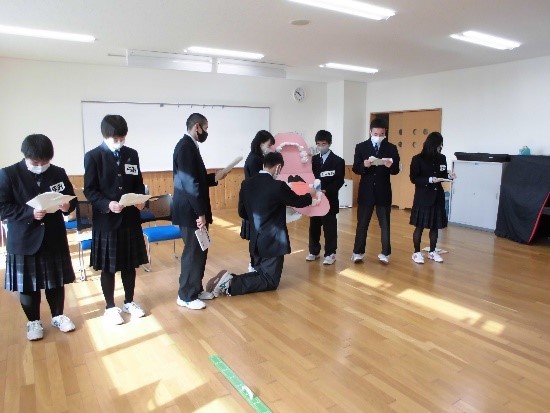 山田高校の保健委員が船越小学校生徒に歯磨き指導を行っている写真。大きな歯の模型を使い六歳臼歯の磨き方のポイントを実演している。